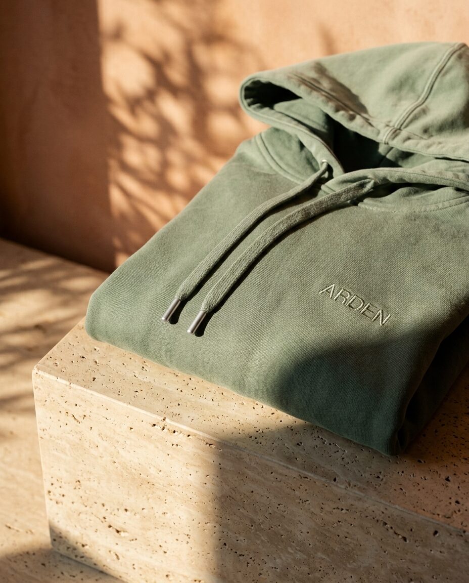The sage hoodie folded on a travertine block, the Arden wordmark on the chest, low sun across it