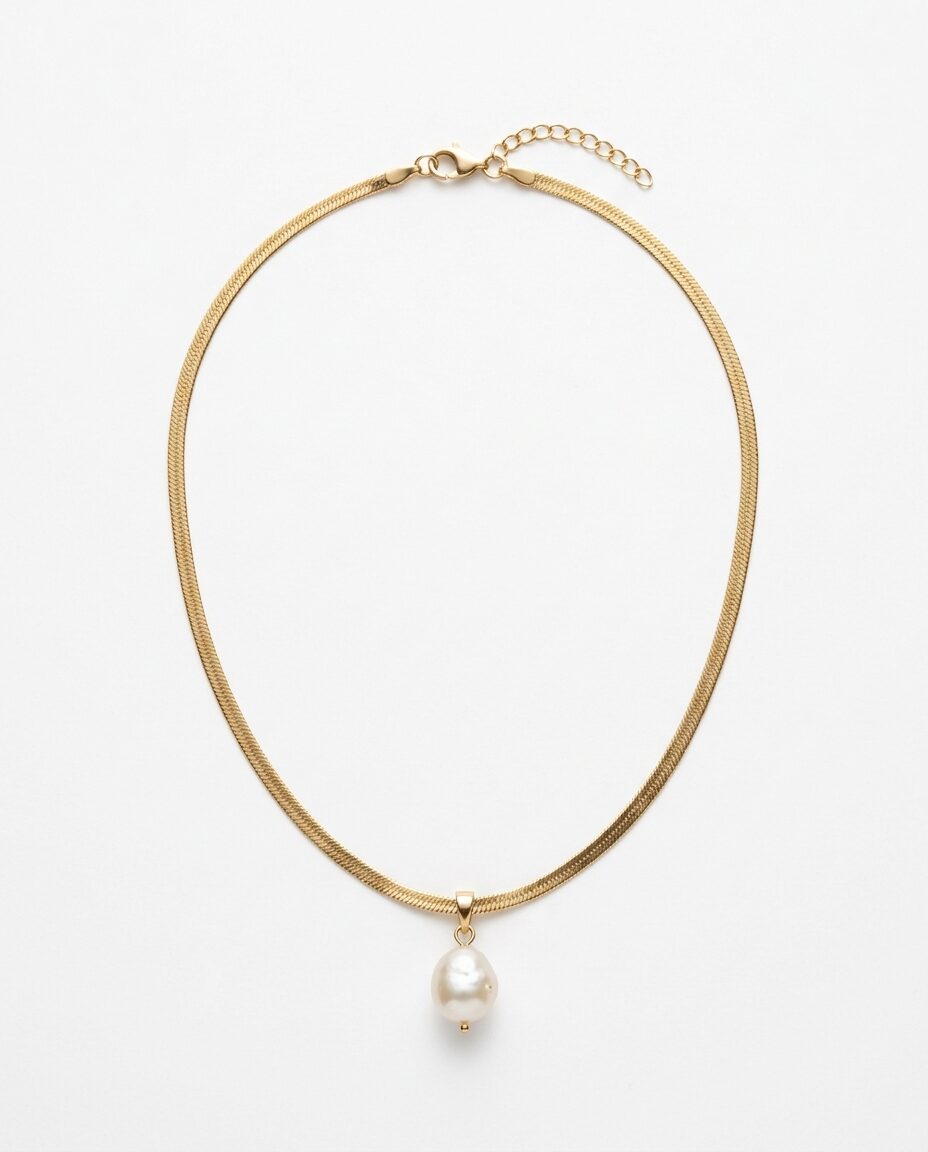 A gold herringbone necklace with a baroque pearl drop, laid flat on white